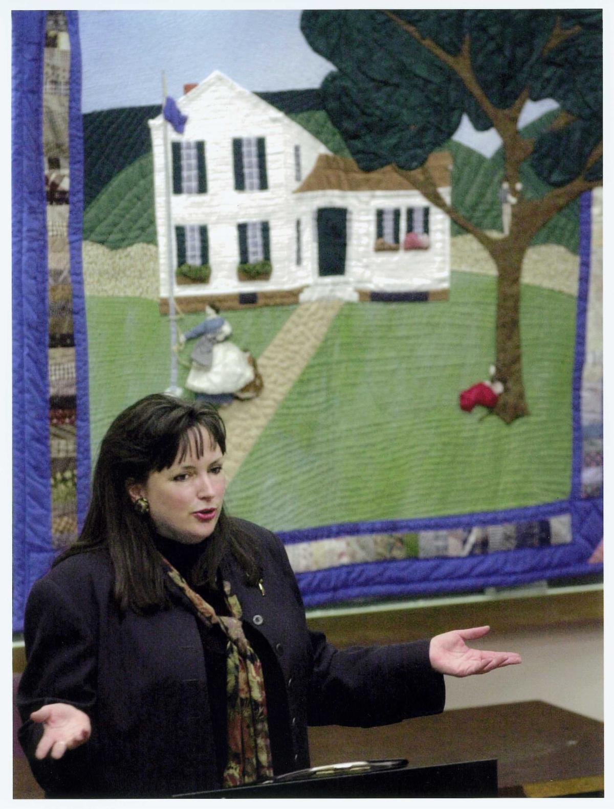 A woman standing in front of a quilt with a white house on it